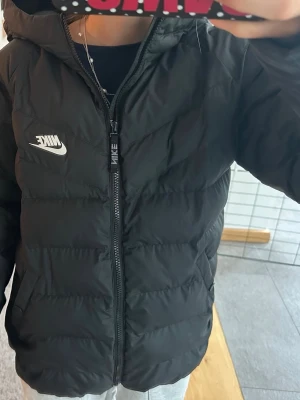 Svart pufferjacka från Nike med huva - Snygg svart pufferjacka från Nike med huva och dragkedja framtill. Jackan har Nike-logga på bröstet och quiltad design som ger en sportig vibe. Perfekt för kalla vinterdagar och riktigt skön att bära. Materialet är lätt och isolerande.