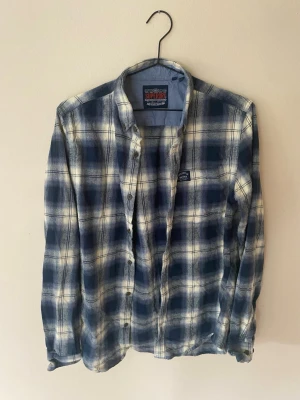 Blåvit rutig skjorta från Superdry - Säljer en blåvit rutig skjorta från Superdry med klassisk krage och knappar framtill. Skjortan har lång ärm och en liten Superdry-logga på bröstet. Perfekt för dig som gillar avslappnad stil och mjuka material. Vintage!