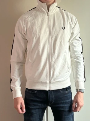 Vit track jacket från Fred Perry - Vit track jacket från Fred Perry med svart rand längs ärmarna och klassisk logga på bröstet. Dragkedja framtill, hög krage och ribbade muddar vid ärmslut och midja. Perfekt för en sportig och stilren look.