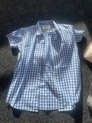 Blårutig kortärmad skjorta Denim Day - Kortärmad skjorta från Denim Day i blåvitt rutigt mönster. Klassisk krage och knäppning framtill. Tillverkad i bomull, perfekt för varma dagar. Snygg och enkel att matcha med jeans eller shorts.