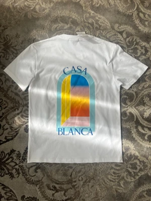 Vit Casa Blanca t-shirt med tryck - Säljer en vit t-shirt från Casa Blanca med färgglatt grafiskt tryck i blått, gult, rosa och orange på både bröstet och ryggen. Klassisk passform och rund halsringning. Perfekt för dig som gillar stilrena men ändå unika plagg.