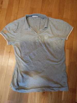 Grå piké t-shirt från sailracing - Snygg grå piké t-shirt från sail racing med korta ärmar och vit broderad logga på bröstet. Krage med knappar framtill och diskreta vita detaljer på ärmslut och krage. Tillverkad i mjuk bomull, perfekt för en avslappnad stil. Det är några hål se bild 5 och 6. Den passar personer runt 165-170 cm lång. Annars riktigt najs tröja. Mvh Rasmus