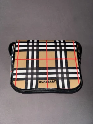 Burberry axelväska rutigt mönster - Snygg axelväska från Burberry med klassiskt rutigt mönster i beige, svart, vitt och röda linjer. Väskan har justerbar svart axelrem och lock med Burberry-logga framtill. Perfekt för dig som gillar stilrena accessoarer med ikonisk design.