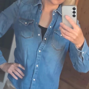Blå jeansskjorta med knappar - Säljer en klassisk blå jeansskjorta med långa ärmar och vita knappar. Skjortan har två bröstfickor med lock och knapp, samt en snygg krage. Använd 1 gång