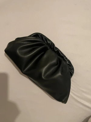 Svart kuvertväska i veganskt läder - Stilren svart kuvertväska i mjukt veganskt läder med puffig, veckad design. Väskan har en minimalistisk look och stängs med magnet. Perfekt för dig som gillar trendiga accessoarer med enkel men cool känsla.