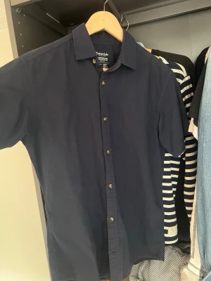 Mörkblå kortärmad skjorta - Snygg mörkblå kortärmad skjorta från Jack & Jones Essentials. Modellen heter The Summer Shirt och har regular collar. Tillverkad i en skön mix av bomull och linne, perfekt för varma dagar. Klassisk knappstängning framtill och stilren design.