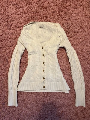 Vit kabelstickad kofta Hollister - Kabelstickad kofta i krämvit färg från Hollister med v-ringning och bruna knappar framtill. Liten broderad logga på bröstet. Långa ärmar och ribbade muddar ger en klassisk look. Perfekt för lager på lager och mysiga dagar.