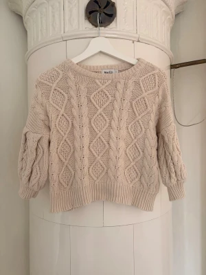 Beige kabelstickad tröja från NA-KD - Säljer en beige kabelstickad tröja från NA-KD med rund halsringning och ballongärmar. Tröjan har ett snyggt flätmönster och ribbade muddar vid ärmslut och nederkant. Cozy vibes.