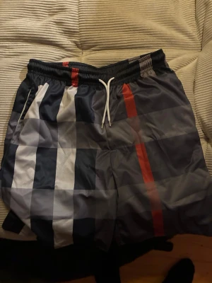 Rutiga shorts med snörning - Snygga shorts med rutigt mönster i svart, grått, vitt och röda detaljer. Shortsen har elastisk midja med vit snörning och är tillverkade i ett lätt syntetmaterial. Perfekta för sommaren och ger en chill vibe.