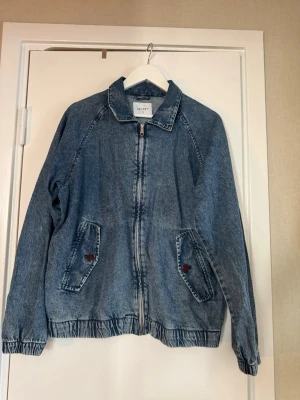 Blå vintage jeansjacka med dragkedja - Blå jeansjacka i klassisk modell med dragkedja framtill och två fickor med knappdetaljer. Jackan har elastisk nederkant och muddar vid ärmsluten för en avslappnad passform.