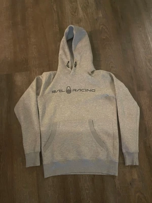 Grå hoodie från Sail Racing - Snygg grå hoodie från Sail Racing med svart logga tryckt på bröstet. Klassisk modell med huva och dragsko samt känguruficka framtill. Perfekt för en avslappnad och sportig stil. Tröjan är i storlek xs men passar mer S/M. Använd 1-2 gånger 