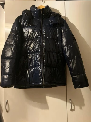 Mörkblå glansig pufferjacka med huva - Säljer en otroligt skön och varm vinter jacka, perfekt till skidbacken eller bara ha till vardagen när det är kallare ute. Helt oanvänd och mycket fint skick, precis som ny. Kom gärna med bud.