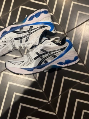 Vita och blå Asics med svart detalj - Säljer ett par snygga Asics i vitt med blå och svarta detaljer. Skorna är använda 2 gånger och  har en sportig design med meshpaneler för bra ventilation och en rejäl sula med blå inslag. Perfekta för dig som gillar streetwear och vill ha något bekvämt och stilrent.