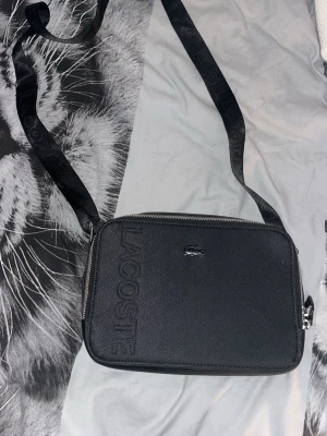 Svart axelväska från Lacoste - Snygg svart axelväska från Lacoste med präglad logga på framsidan och justerbar axelrem med Lacoste-tryck. Väskan har en stilren, rektangulär form och är tillverkad i slitstarkt material med dragkedja upptill. Perfekt för att bära det viktigaste.