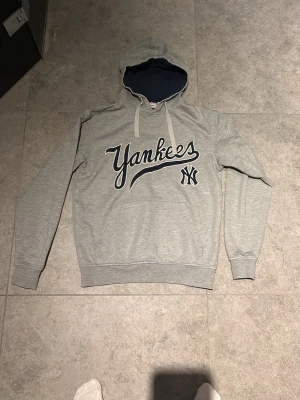 Grå Yankees hoodie med tryck - Säljer en grå hoodie med stor Yankees-logga och text på bröstet. Tröjan har huva med mörkblått foder och vita dragsnören. Klassisk passform med ribbade muddar vid ärmar och nederkant. Perfekt för dig som gillar sportig streetstyle.