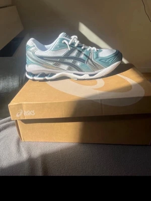 Asics vita och turkosa sneakers - Snygga sneakers från Asics i vitt och turkost med silverdetaljer. Skorna har en sportig design med meshpaneler för bra ventilation och klassiska Asics-ränder på sidan. Yttersulan är vit och ger bra grepp. Perfekta för dig som gillar streetwear och sportig stil.