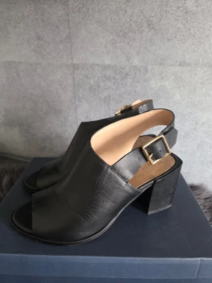 Novita svarta skinnpumps peeptoe slingback – stl 38 - 🖤 Stilrena svarta pumps från Novita i skinn. Modell med öppen tå, bred stabil blockklack och justerbar rem runt hälen med guldfärgat spänne.  🔖 Stl 38 🧵 Skinn 📦 Originalkartong medföljer 🏷️ Nypris 999 kr  ✨ Mycket fint skick. Normala användningsveck i skinnet samt ytliga märken på klackarna och mindre bruksspår vid sulkanterna förekommer. Se bilder för egen bedömning.