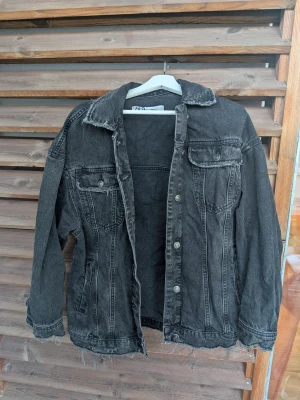 Svart tvättad jeansjacka - Snygg svart tvättad jeansjacka med klassisk krage, två bröstfickor med lock och knappar samt slitna kanter nedtill. Jackan har en avslappnad passform och tydliga sömmar som ger en cool vintagekänsla. Perfekt för dig som gillar streetstyle.