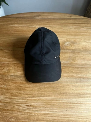 Svart keps från Nike med logga - Svart keps från Nike med klassisk böjd skärm och silverfärgad Swoosh-logga på sidan. Kepsen är i ett mjukt tygmaterial och har en stilren, sportigpp look som passar till det mesta.