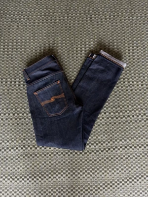 Nudie jeans dry Japan selvage - Grim Tim - Säljer ett par Nudie Jeans Grim Tim Dry Japan Selvage i storlek W30/L32.  Slim fit med rakare ben och tillverkade i japansk selvedge-denim från Kaihara Mills. Mörk indigo/raw denim med en rigid känsla och tydlig twillstruktur. Ett riktigt snyggt par jeans för dig som gillar klassisk denim, selvedge och en stilren passform.  Storlek: W30/L32 Modell: Grim Tim Färg: Mörk indigo Märke: Nudie Jeans Material: Japanese selvedge denim