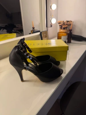 Svarta pumps med gulddetaljer - Säljer ett par svarta pumps med öppen tå och höga klackar. Skorna har tre remmar med guldfärgade fyrkantiga detaljer över foten och en elegant siluett. Materialet ser ut att vara konstläder med lätt struktur. Banden med nitar går att ta av om man vill ha en simplare stil.