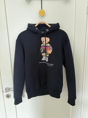 Svart hoodie med Polo Bear-tryck - Svart hoodie från Polo Ralph Lauren med ikoniskt Polo Bear-tryck på bröstet. Hoodien har dragsko i huvan, stor magficka och ribbade muddar. Perfekt för en chill och stilren look.
