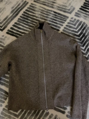Beige ribbstickad kofta med dragkedja - Säljer en beige ribbstickad kofta med hög krage och hel dragkedja framtill. Koftan har långa ärmar och en rak passform som ger en avslappnad look. Perfekt att slänga på sig när det är kyligt ute.