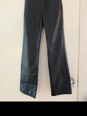 Svarta skinn byxor med raka ben - Säljer ett par svarta skinnbyxor med raka ben och medel hög midja. Byxorna har en glansig finish som ger en cool look och två bakfickor. Perfekta för dig som vill ha ett par trendiga byxor med lite edge. Aldrig använda 