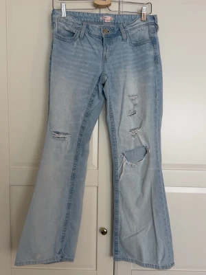 Ljusblå slitna bootcut jeans - Säljer ett par ljusblå bootcut jeans med flera slitningar och hål på benen för en trendig look. Jeansen har klassisk femficksmodell, hög midja och är tillverkade i denim. Perfekta för dig som gillar en avslappnad och cool stil.
