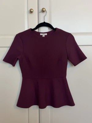 Vinröd peplumblus från H&M - Snygg vinröd peplumblus från H&M med rund halsringning och korta ärmar. Blusen har en figurnära passform upptill och en utsvängd nederdel som ger en feminin siluett. Tillverkad i ett stretchigt material som sitter bekvämt.