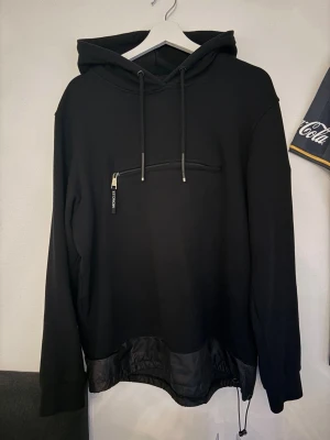 Svart hoodie med dragkedja och snörning - Säljer en svart hoodie med huva och dragsko. Framtill finns en cool dragkedja över bröstet och nederkanten har ett snyggt tygparti med snörning. Hoodien har långa ärmar och en avslappnad passform, perfekt för en streetwear-look.