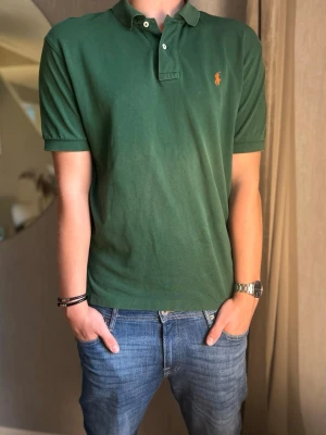 Grön pikétröja från Ralph Lauren - Snygg grön pikétröja från Ralph Lauren med klassisk krage och korta ärmar. Tröjan har den ikoniska broderade loggan på bröstet och är tillverkad i mjuk bomull. Perfekt för en avslappnad men stilren look. Modellen är 185cm lång och den är perfekt i längden men lite liten på de andra ställena 