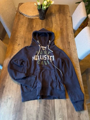 Vintage Mörkblå hoodie från Hollister - Mörkblå hoodie från Hollister med stor magficka och vit dragsko i huvan. Tryckt text 'Hollister Cali' i vitt och grönt på bröstet. Klassisk relaxed fit och mjukt material, perfekt för chill dagar.