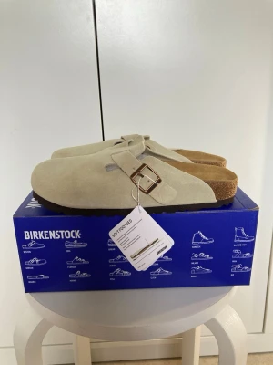 Beige Birkenstock tofflor mocka - Snygga beige tofflor från Birkenstock i mjuk mocka med klassisk korksula och justerbart spänne i metall. Perfekta för dig som gillar stilren och bekväm design. Innersulan är formad för extra komfort och tofflorna har en tidlös look.