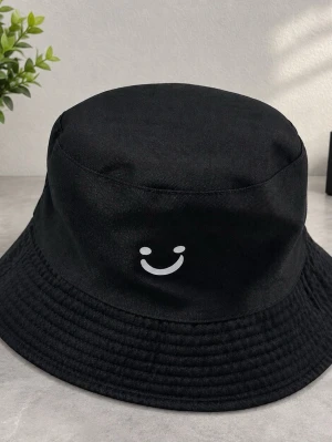 Svart bucket hat med smiley - Cool svart bucket hat i bomull med en vit smiley framtill. Insidan har ett unikt svartvitt koskinnsmönster som ger hatten extra edge. Perfekt accessoar för dig som vill sticka ut och addera lite personlighet till din stil.