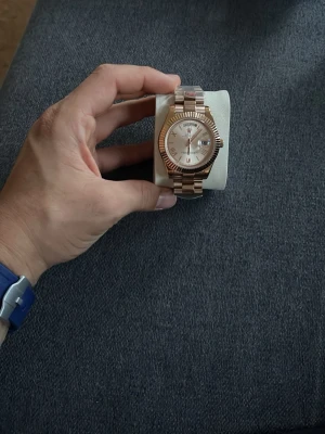 Roséguld klocka med metallarmband - Snygg klocka i roséguld med metallarmband och räfflad bezel. Urtavlan är ljus med romerska siffror och datumvisning. Klockan har en klassisk och elegant design som passar till många olika stilar.