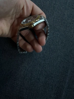 Stilren klocka i silver och guld - Snygg klocka med tvåfärgad länk i silver och guld samt svart urtavla. Boetten har guldfärgad räfflad kant och länken är i metall. Perfekt accessoar för dig som vill ha en klassisk men ändå modern look.