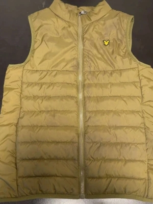 Olivgrön dunväst med dragkedja - Snygg olivgrön dunväst med quiltad design och hög krage. Västen har en gul logga på bröstet och stängs med dragkedja framtill. Perfekt att slänga över en hoodie eller tröja för extra värme. Materialet är glansigt och lätt, vilket gör den enkel att bära.