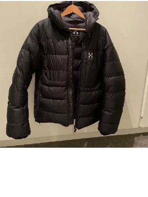 Svart dunjacka från Haglöfs - Säljer en svart dunjacka från Haglöfs med huva och dragkedja framtill. Jackan har en quiltad design, diskret Haglöfs-logga på bröstet och praktiska sidofickor. Perfekt för kalla vinterdagar och riktigt varm tack vare dunfyllningen. Har litet hål som är sytt men inget som syns. Varm och skön 