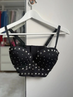 Svart bustier med nitar - Säljer en svart bustier med justerbara axelband och vadderade kupor. Bustiern är dekorerad med silvriga nitar i olika storlekar och har en edgy, rockig look. Materialet ser ut att vara en blandning av polyester och elastan för en tajt passform.