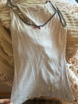Odd Molly linne med rosett - Säljer en ribbad ljusgrå linnetopp med tunna axelband och en liten lila rosett framtill. Toppen är figurnära och har en mjuk, stretchig känsla. Perfekt att styla med jeans eller kjol för en enkel och snygg look.