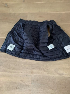 Mörkblå dunväst från Moncler - Snygg mörkblå dunväst från Moncler med quiltad design, ståkrage och dragkedja med röd, vit och blå rand. Två fickor med tryckknappar och Moncler-logga på bröstet. Perfekt för lager på lager och streetstyle.