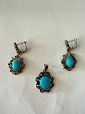 Silver örhängen och hängsmycke med turkosa stenar - Säljer ett par vintageinspirerade örhängen i äkta silver (925)  med ovala turkosa stenar i mitten. Örhängena har en dekorativ ram med små pärldetaljer runt stenen och klassisk låsning. Hängsmycket matchar örhängena och har samma turkosa sten och detaljer. Perfekt för dig som gillar boho eller retrostil. Aldrig använda. 