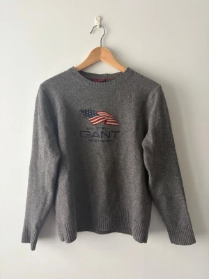 GANT flaggtröja i 100% lammull - Klassisk grå stickad tröja från GANT med den ikoniska amerikanska flaggan och "EST. 1949 NEW HAVEN" broderat på bröstet. Säljs inte längre i butik (nypris för motsvarande modell ca 2 199 kr), så detta är ett riktigt vintagefynd! Tillverkad i varm och skön 100% lammull (Lambswool). Storlek M men passar även S.