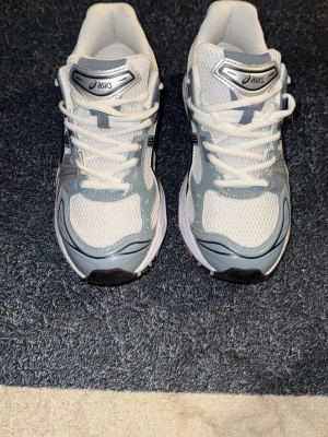 Asics vita och grå sneakers - Snygga sneakers från Asics i vitt och ljusgrått med klassiska svarta detaljer på sidorna. Ovandelen är i mesh och syntetmaterial för bra andningsförmåga och komfort. Skorna har en sportig look med lågt skaft och vita snören.