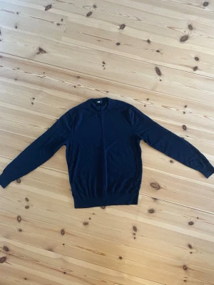 Mörkblå stickad tröja från Uniqlo - En enkel och stilren mörkblå stickad tröja med rund halsringning och långa ärmar. Tröjan har ribbade muddar vid ärmslut och nederkant. Perfekt att matcha med jeans eller kjol för en clean look. 100% ull