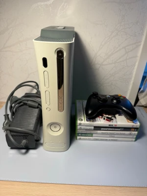 Xbox 360 med handkontroll och spel - Säljer en vit Xbox 360 med svart trådlös handkontroll, strömadapter och fem spel: NHL 10, Grand Theft Auto V, Need for Speed Hot Pursuit, FIFA 10 och Street Fighter X Tekken. Konsolen har en stilren design i plast och metall.