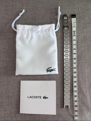 Lacoste armband - Säljer en snygg armbandslänk från Lacoste i silverfärgad metall. Länken har ett stilrent mönster på varje länk och klassisk Lacoste-logga på spännet. Perfekt för dig som vill uppgradera din klocka med en exklusiv känsla.
