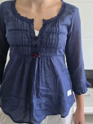 Odd molly Blå blus med spetsdetaljer - Söt blå blus med trekvartsärmar och spetskantad v-ringning. Blusen har dekorativa veck och spetsband framtill samt en liten rosett under bysten. Tillverkad i ett tunt, lätt material som ger ett luftigt intryck. Volangdetaljer vid ärmslut och lös passform.