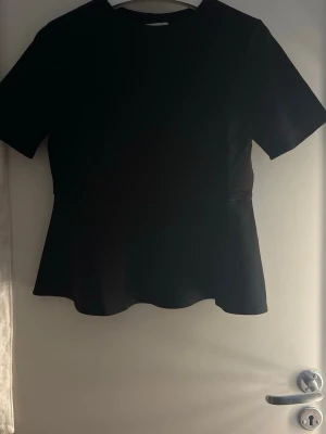 Svart topp med kort ärm - En stilren svart topp med rund halsringning och korta ärmar. Toppen har en lätt utsvängd nederkant som ger en snygg siluett. Perfekt att matcha med både jeans och kjol för en clean look. Säljer pågrund av fel storlek så har ej använts